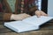 A closed book or notebook lies on a wooden table. Sitting next to a woman