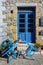 Closed sidewalk cafe with chairs tilted onto small bistro table in front of brightly painted doors in old rock building