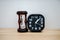 Closeup black alarm clock for decorate next to hourglass, wooden desk front concrete wall background.