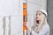 Closeup blonde girl designer foreman in white construction helmet measures the grey wall osibit orange building level in house