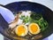 Closeup of a Bowl of Ramen with Two Eggs, Chicken, Greens and Green Onion. White Plastic spoon, black ceramic bowl, sesame seeds