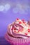 Closeup cupcake with creamy pink and white top decorated with little hearts on purple bokeh background.