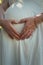 Closeup cute composition with a pregnant young woman wearing white dress makes a heart shaped symbol with her hands on her belly.