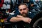 Closeup face of bearded cycling repairman standing leaning on bicycle in repair bike workshop with dark interior