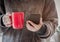 Closeup of female hands with a red mug coffee beverage. Beautiful girl in grey sweater holding cup of tea or coffee and smartphone