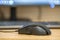 Closeup of a gaming black mouse controller on an empty office desk with computer screen on blurred background