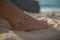 Closeup of girl right foot with boho style bracelet on sand, model sits on beach