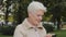 Closeup gray-haired Mature woman in casual clothes walking outdoors in city park uses cellphone modern gadget smartphone