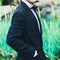 Closeup groom in a blue suit, white shirt and tie butterfly holds hands in pockets on a background of a green bush