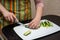 Closeup hands girl in a striped apron in the kitchen is slicing cucumbers into strips on a whiteBoard on the black table