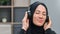 Closeup happy face of Muslim woman in black headscarf listening music in headphones relaxing at home