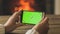 Closeup image of young woman holding smartphone and making image of burning fireplace. Empty green screen for inserting