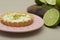 Closeup of a key lime tartlet on a pink plate with key lime fruits on a wooden board