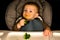 Closeup of male baby sitting in high chair holding broccoli brown sauce on mouth black background