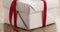 Closeup pan of white paper gift box with thin red ribbon bow on old wood table