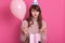 Closeup portrait of beautiful disappointed girl with gift package and balloon, wearing pink sweater and birthday hat, posing