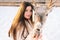 Closeup portrait of beautiful young woman wearing fur coat stroking and feeding cute young reindeer on snowy deer farm