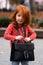 Closeup portrait of cute adorable smiling little red-haired Caucasian girl child standing with mama`s big bag in autumn