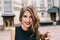 Closeup portrait of effective girl with long curly hair smiling to camera on street on building background. She wears