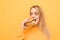 Closeup portrait of a girl biting a burger with her eyes closed on a yellow background, wearing orange clothing.Hungry girl eats a