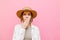 Closeup portrait of shocked girl in hat on pink background, looking into camera with mouth covered with hand and surprised look.