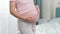 Closeup of pregnant woman in pajamas standing at big window and stroking big belly. Concept of happiness during