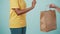 Closeup shot. Delivery woman in yellow uniform giving female client order paper bag with products. Isolated on blue