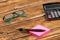 Closeup shot of a pen on pink stickers with glasses and a calculator on a wooden table