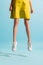 Closeup slender female legs in summer dress and white sneakers hover above the floor isolated on blue background