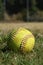 Closeup of a used yellow softball with red seams sitting in grass.