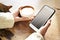 Closeup view woman hands using mobile smartphone and cup of coffee. Space mockup for design layout. Sunlight, slab wooden table.