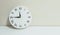 Closeup white clock for decorate show a quarter to twelve or 11:45 a.m. on white wood desk and cream wallpaper textured background