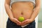 Closeup womans stomach with unzipped jeans and shirt lifted up, holding apple between hands, underwear visible