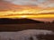 A cloudy daybreak above inversion and hills. Dark peaks of trees make contrast with yellow and orange clouds on the sky, last snow