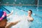 Coach In Water Gives Children Swimming Lesson As They Sit On Edge Of Pool And Splash Him