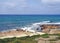 Coastal pathway leading to a white bench with sunlit waves breaking on rocks and a bright blue sea in paphos cyprus