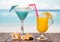 Cocktail and glass of orange juice with umbrellas, straws, starfish, smooth stone and sea shell on background of sea.
