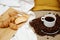 coffee beans sprinkled on a white saucer from above a cup with coffee. next to a croissant on a wooden board