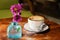 Coffee cup with cappuccino and pink flowers in bottle on the worn polished tabletop side view