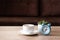 Coffee cup, flower pot and clock on wooden table against defocused sofa with pillows. Front view. Good morning concept. Mock-up