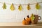 Coffee cup and pumpkin decoration on wooden shelf over brick wall background with autumn leaf garland. Thanksgiving holiday