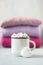 Coffee with marshmallows in white enameled metal cup and white heart on the background of stack of pink knitted sweaters on table