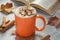 Coffee with whipped cream and marshmallow in bright orange mug on grey table. An open book in soft focus in background. Fall time