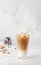 Cold coffee with milk and ice cubes in a tall glass with bamboo straw, light grey background.