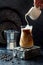 Cold refreshing iced coffee in a tall glass and coffee beans on dark background. Pouring cream into glass with iced coffee.