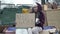 Cold, unkempt-looking young woman sits with a begging cup by a pile of rubbish and holds up a handwritten NEED WORK