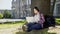 College student sitting under tree in campus, using laptop, writing final paper