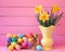 Colorful Easter Eggs in Carton with Vintage Yellow Vase filled with Spring Daffodils against Bright Pink Wood Board Background wit