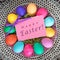 Colorful Easter Eggs on a Plate in center with Pink Happy Easter Name Tag Card on Vintage Lace Tablecloth. A Square crop with goo