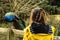 Colorful male peacock on a wooden fence. Young slim teenager girl in yellow jacket looking at the bird, back to viewer. Scene in a